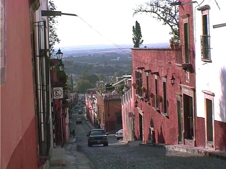 Scenic street view looking down a long slope with building close to kerb on both sides, and countryside fading away in the distance.