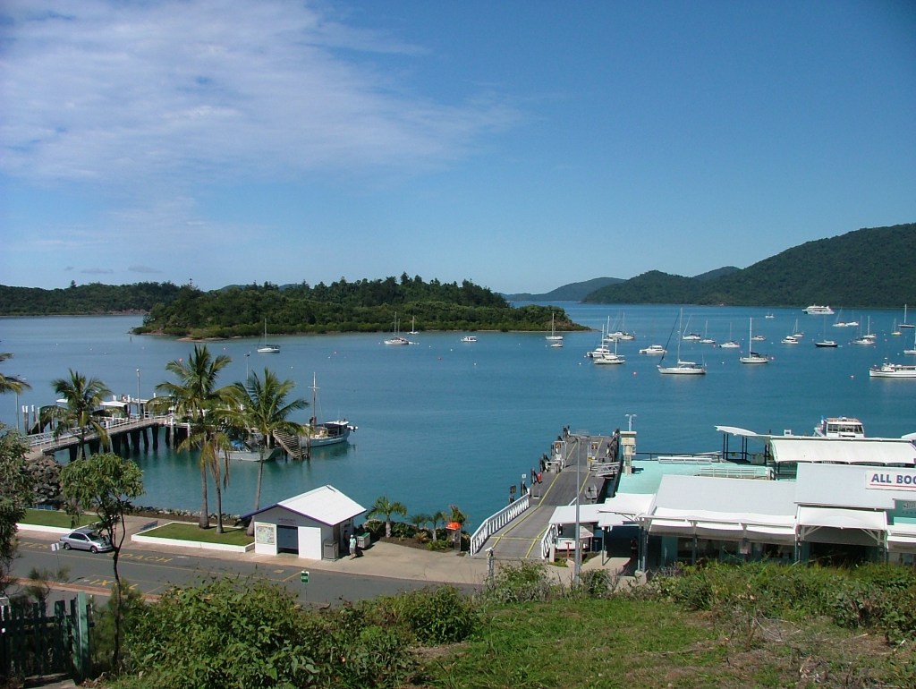 Picturesque scene overlooking building and large jetty on shore next to large tropical bay with lots of moored boats and an island.