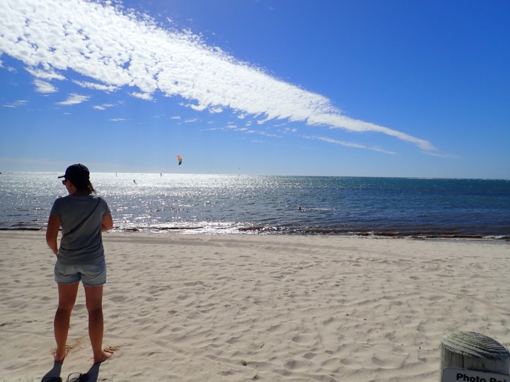 Picturesque Beach scene with white sand, looking seawards with sunlight sparkling on the water and sailboarders in the distance.  Woman standing towards the ocean.